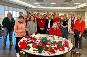 A large group of knitters posed around a table of hats they knitted that were donated.