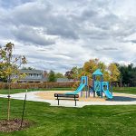 View of a playground, park bench, sidewalk and new tree planting.