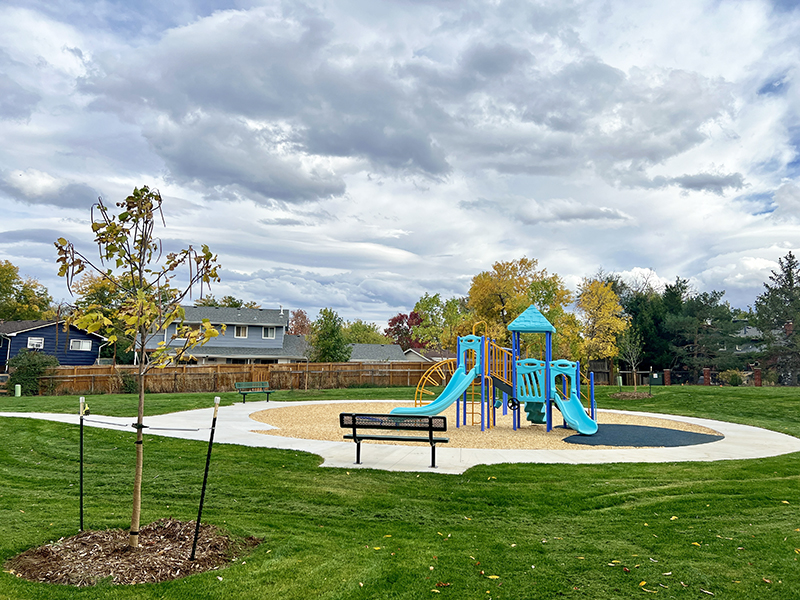 View of a playground, park bench, sidewalk and new tree planting.