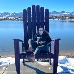 Woman sitting in an oversized purple adirondack chair near a body of water with mountains in the background.