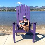 Woman and child sitting in an oversized purple adirondack chair near a body of water with mountains in the background.