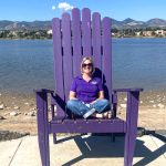 Woman sitting in an oversized purple adirondack chair near a body of water with mountains in the background.