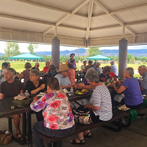 A group of people sitting under a park shelter eating at picnic tables.