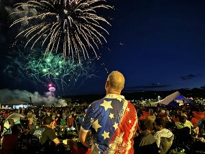 Man in a patriotic shirt standing amongst a crowd of people watching a firework display.